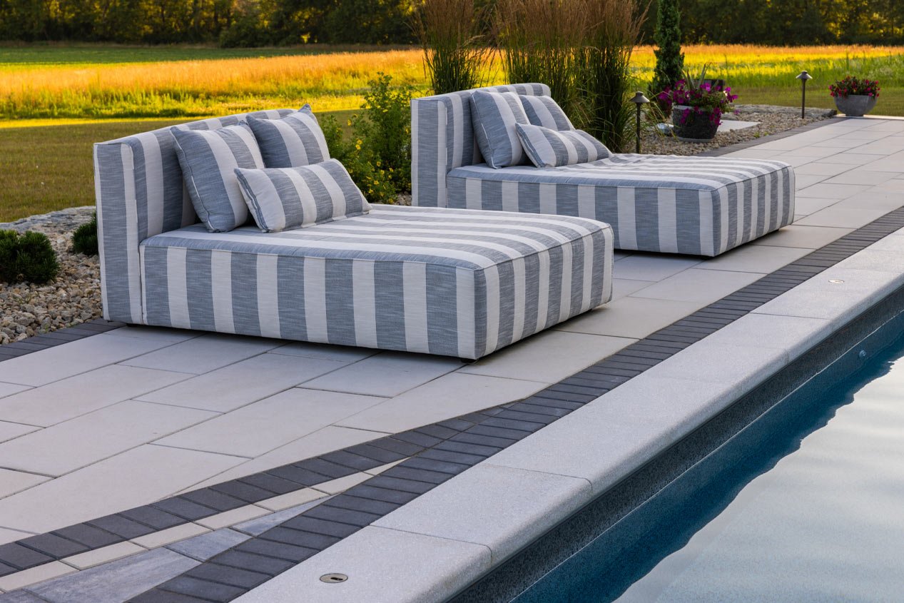 Gray-and-white striped comfy chaise lounges sit on a concrete paver pool deck that includes oversized warm white pavers, bright salt-colored pool coping pavers, and a charcoal-colored border around the edge of the pool.