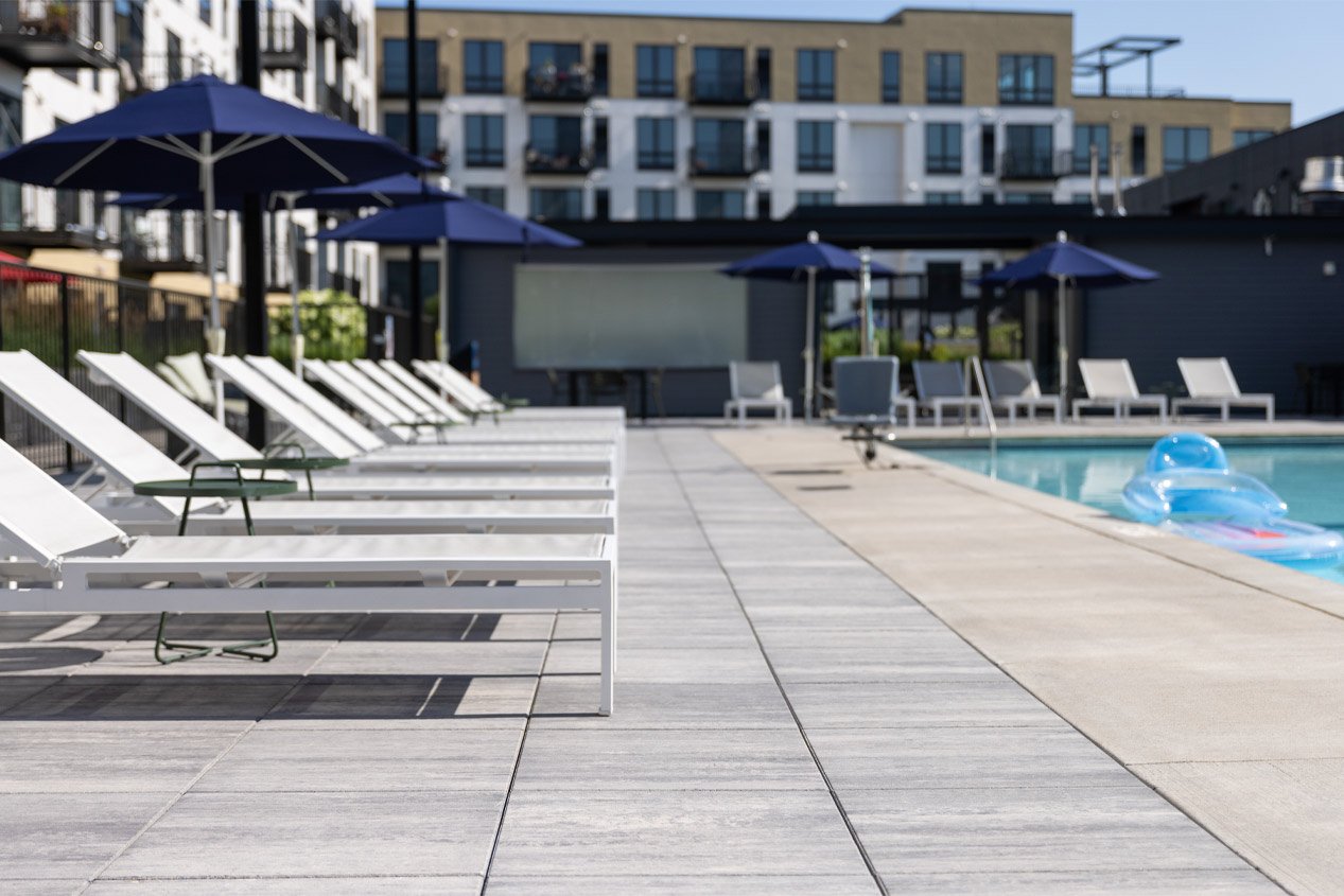 A resort-style swimming pool at an apartment complex has a pool deck made with warm white and grey blend concrete deck pavers, which are installed on pedestals to promote drainage.