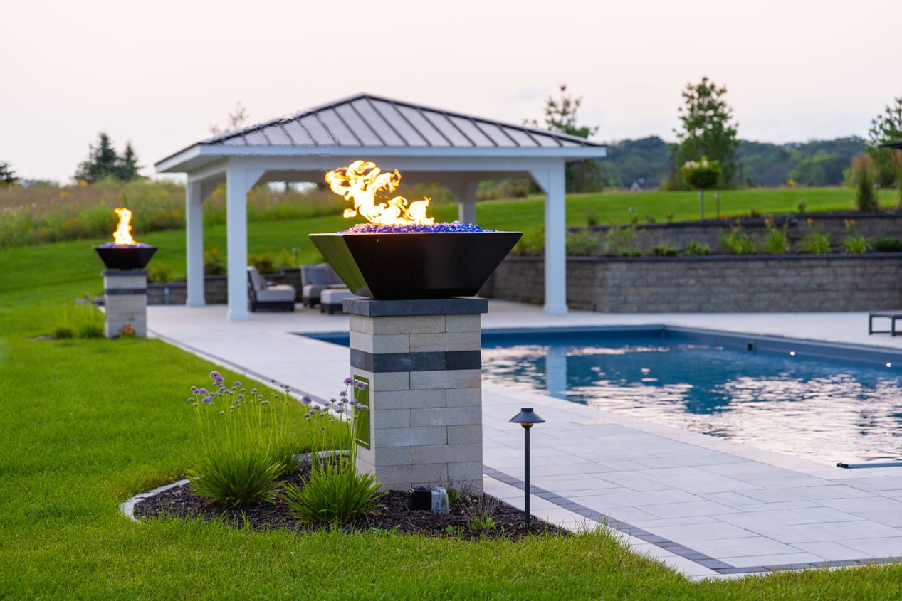 Next to a serene rectangular swimming pool in a lush green backyard, a warm white column built with concrete retaining wall blocks supports a gas fire feature.