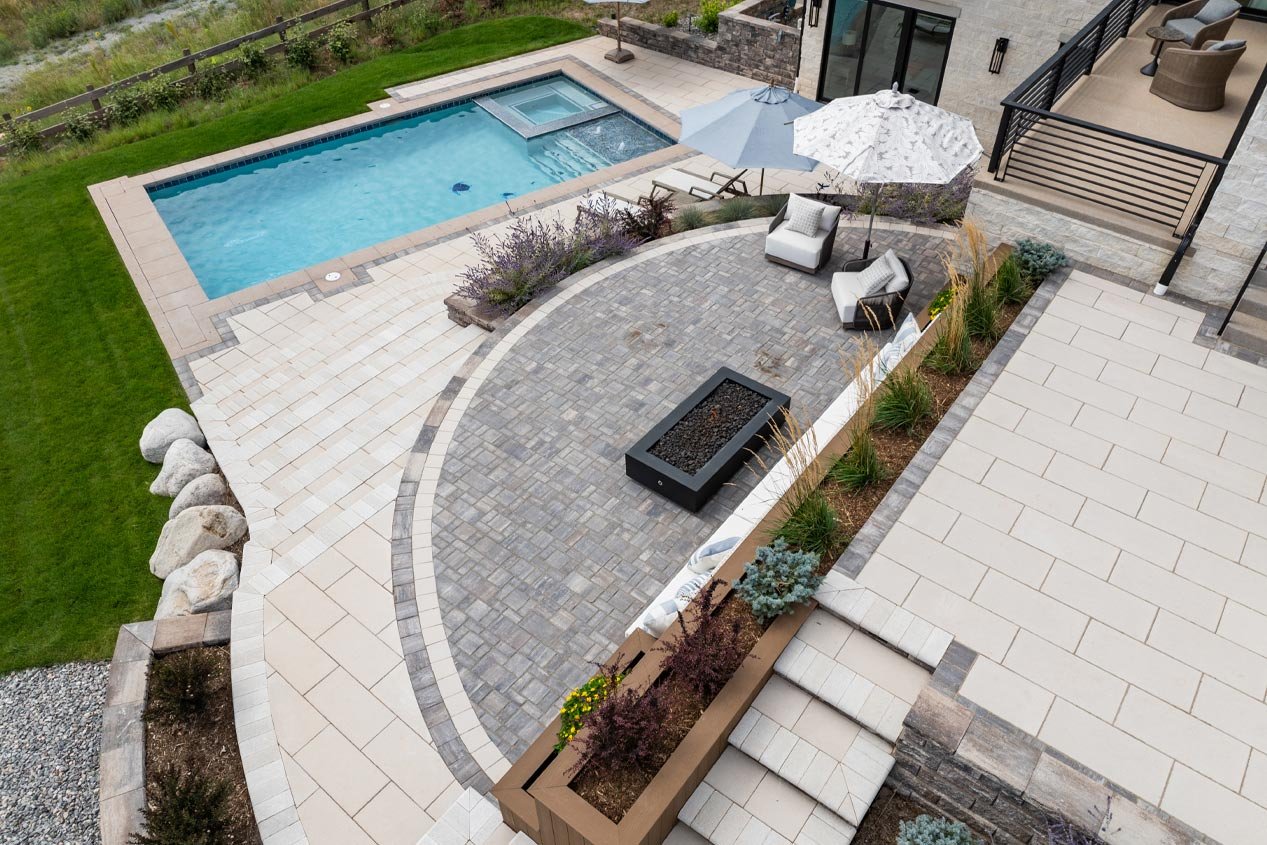 A multi-tiered patio and pool deck with beige concrete pavers includes a semi-circular patio tier elevated above the pool deck, paved with permeable pavers in a brown, gray and warm white blend to promote drainage.