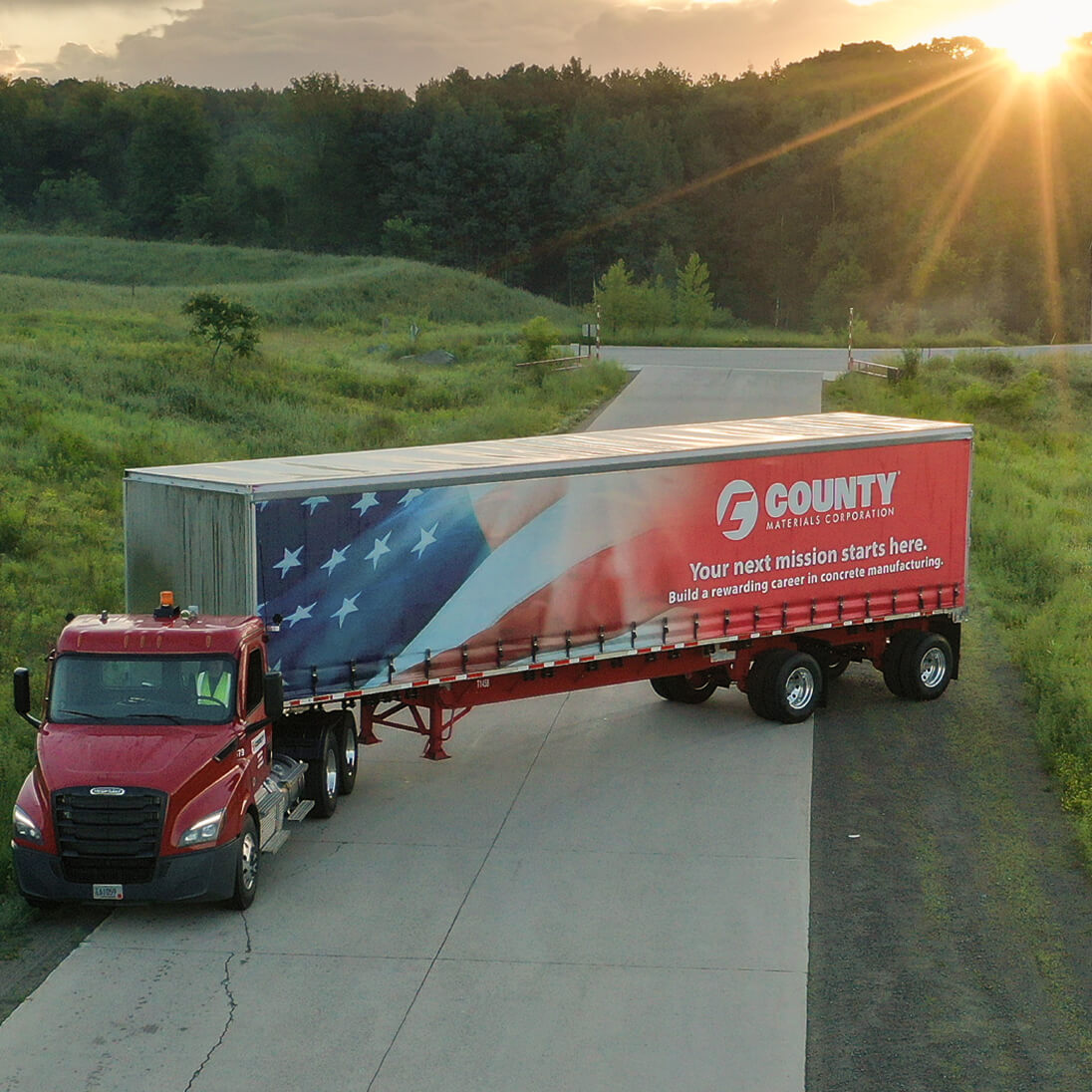 County Materials semi truck hauling an American flag–branded trailer on a rural roadway at sunset