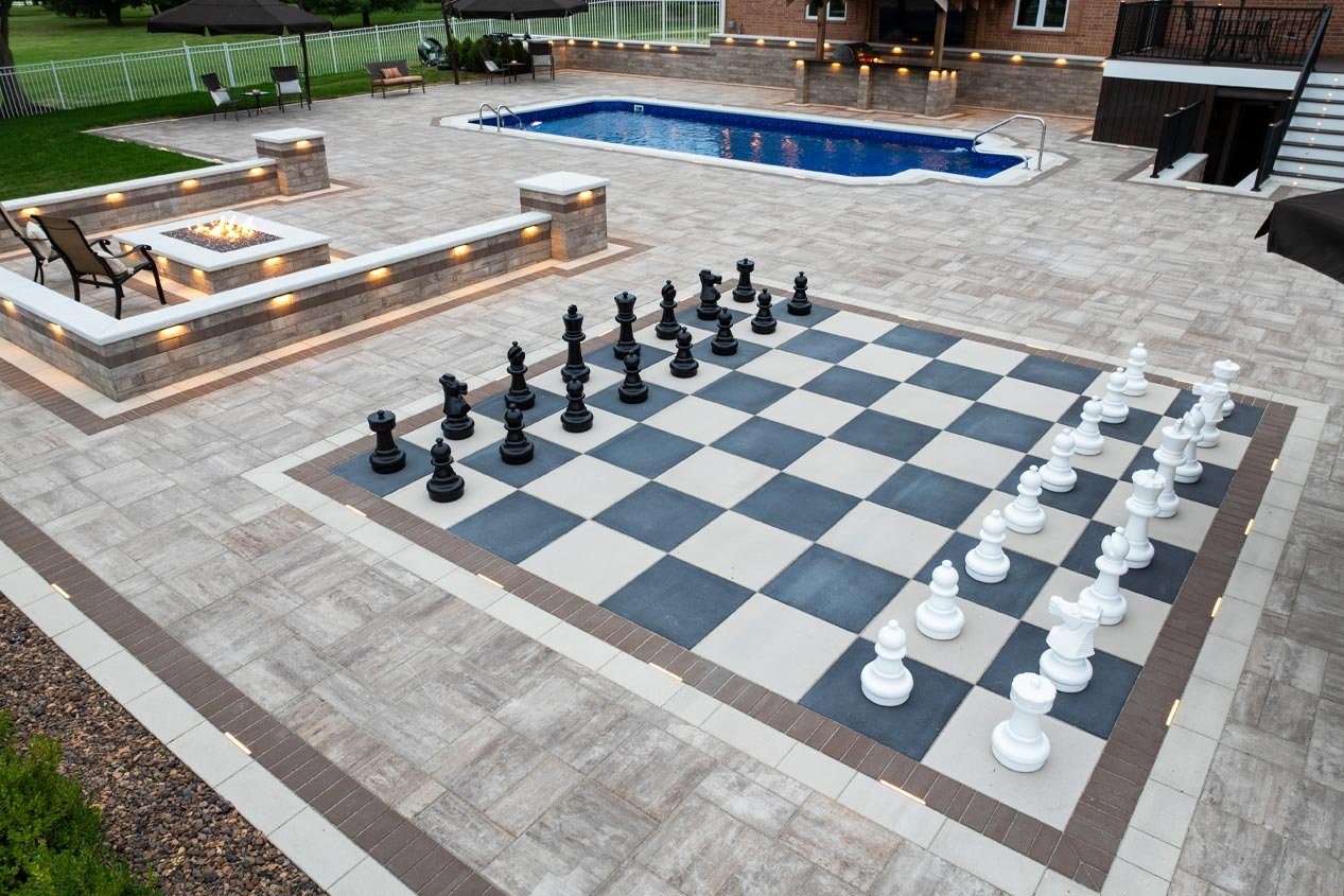 An expansive concrete paver pool deck that includes a giant chess board made with black and white concrete deck pavers and an intimate seating area around a fire feature made with concrete wall blocks.