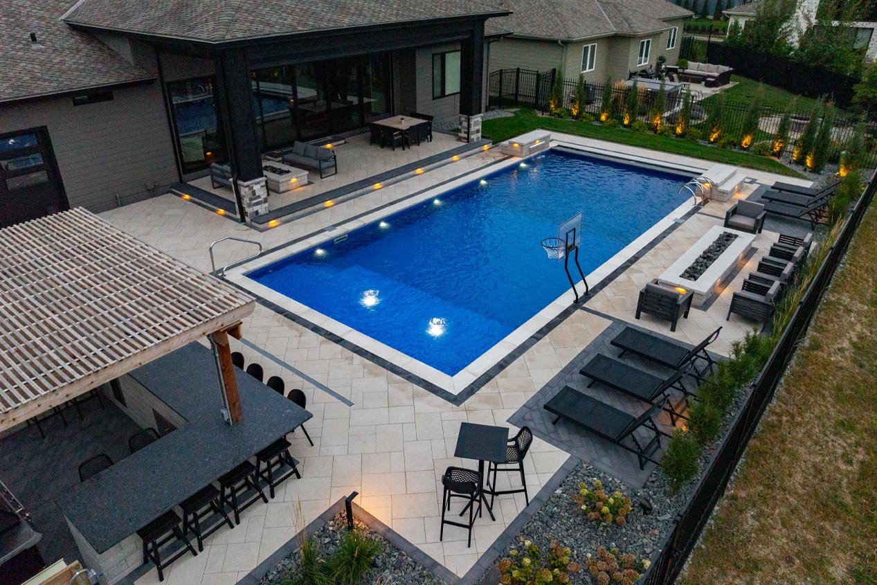 A high-end backyard pool deck with warm white concrete pavers and a charcoal border and accents includes many seating areas and an outdoor bar area under a shade pergola.