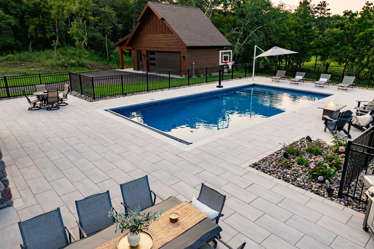A warm white and gray blend concrete paver pool deck is surrounded by an elegant black metal fence and low-maintenance gravel gardens.