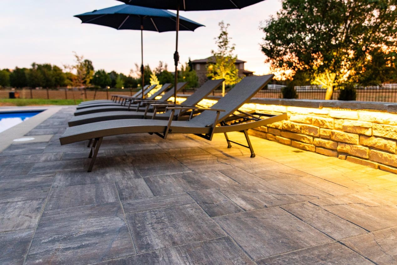 Lighting features built into a low seat wall illuminate a lounge chair area on a concrete paver pool deck at dusk.