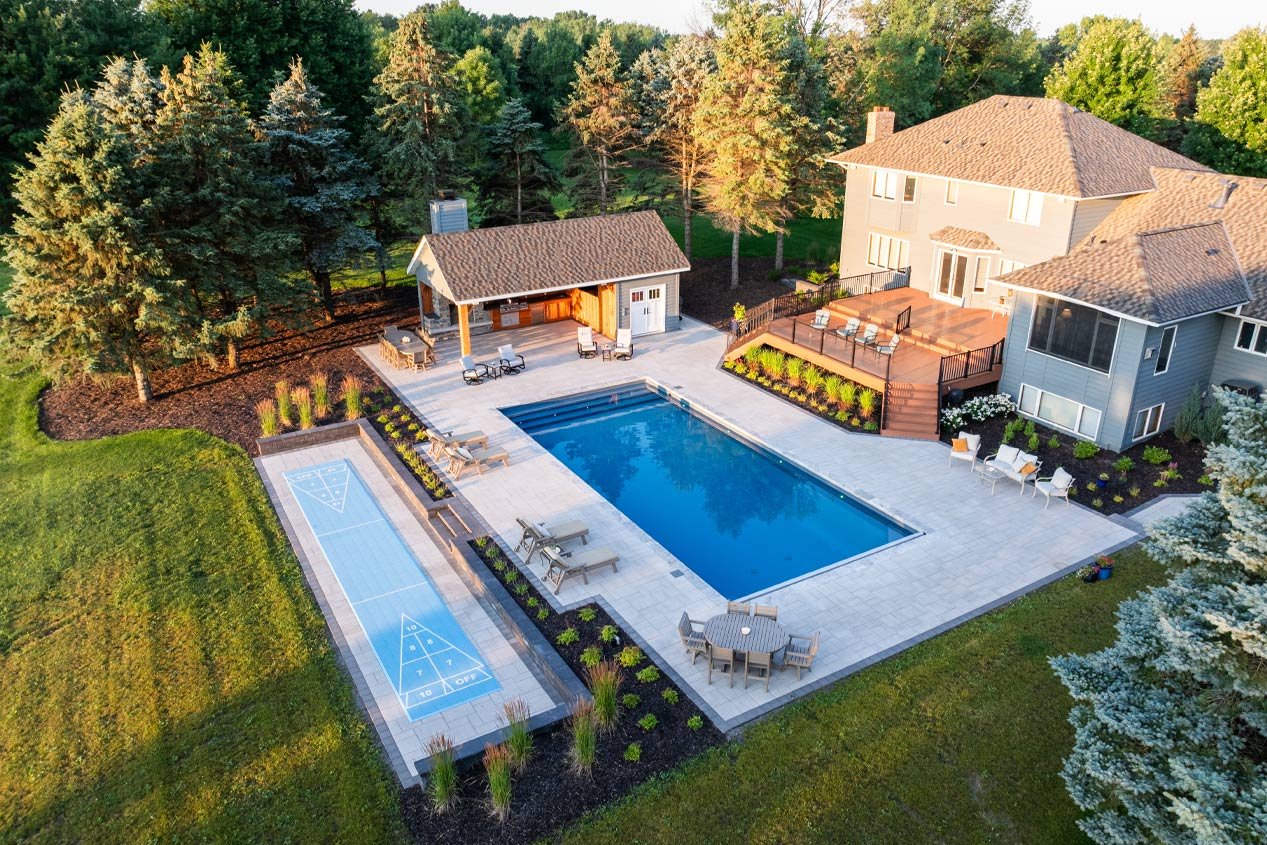 An overhead view of a backyard swimming pool deck at sunset that includes a lower-tier rectangular section for a shuffleboard court.