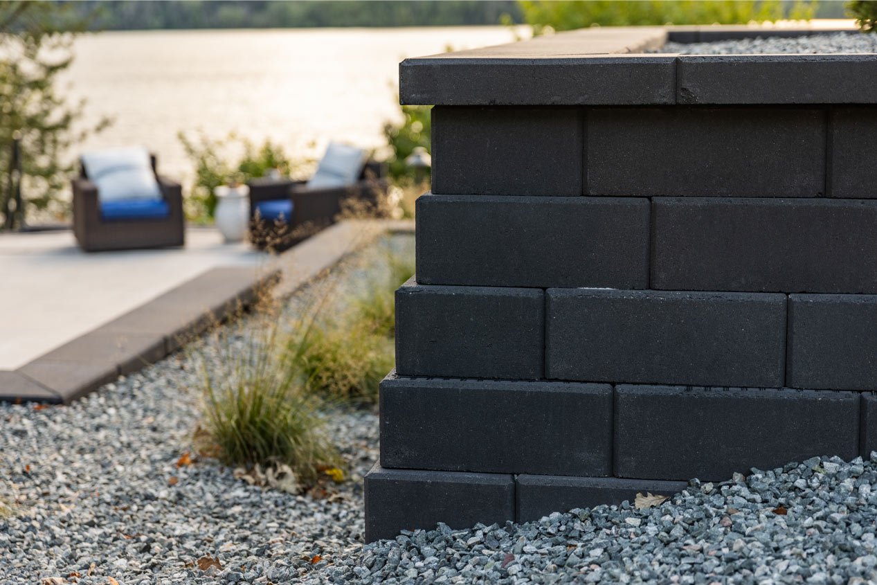 A close-up shot of an outside corner of a dark gray retaining wall with two lounge chairs in soft focus in the background.