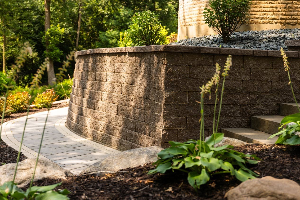 A walkway curves around a Tribute Splitface retaining wall and leads into a staircase in a lush, shaded backyard.