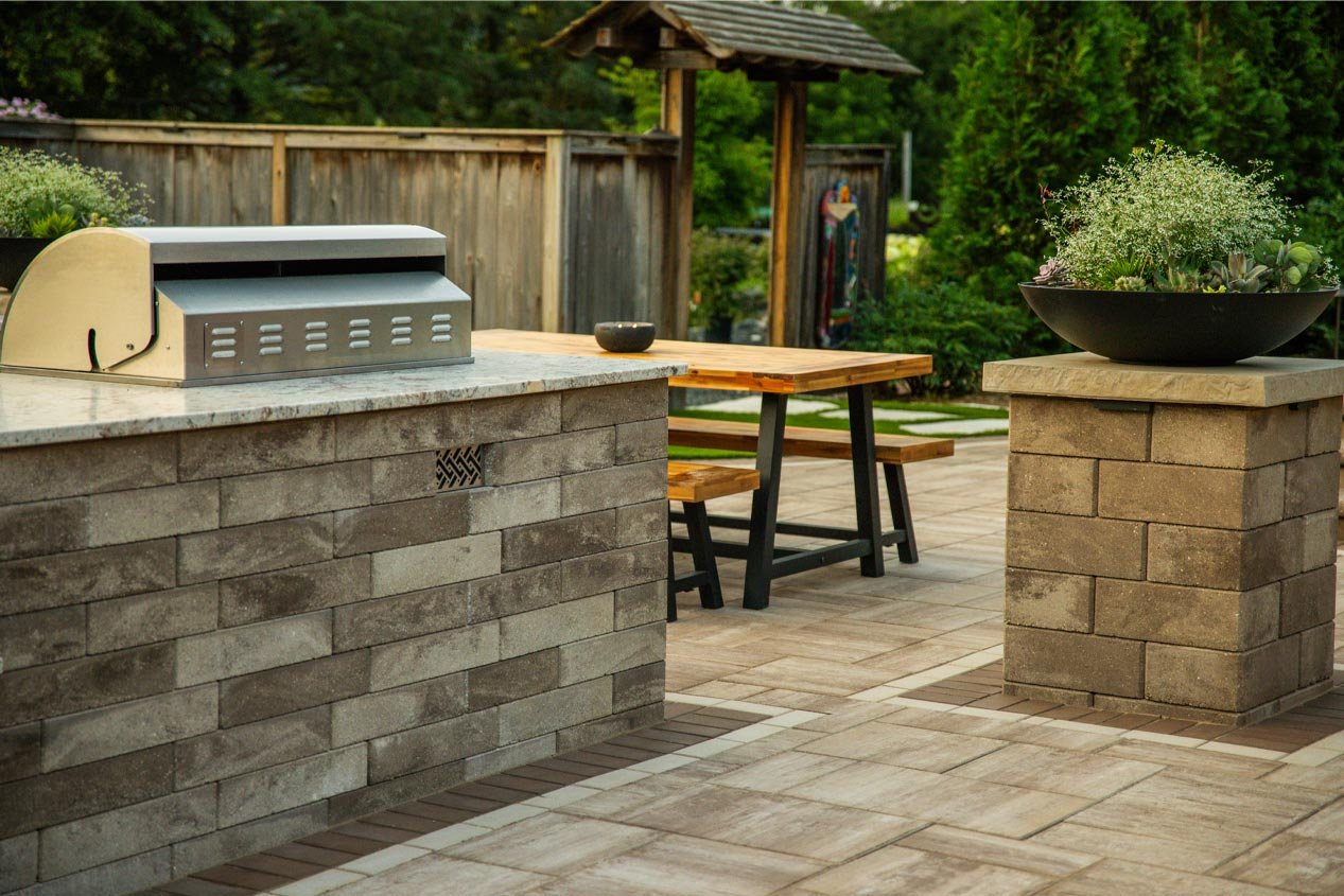 A bar made with concrete landscape units with a built-in gas grill sits on a patio with a picnic table and decorative columns and planters.