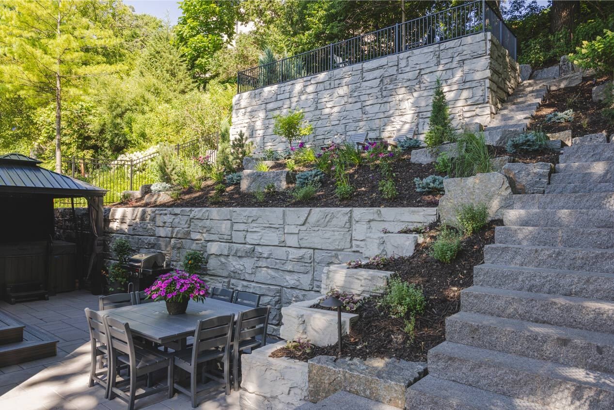 A tall hillside is retained with two tiers of precast modular block retaining walls, with a staircase leading from a lower patio to the top of the walls.