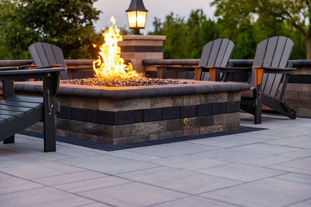 A blazing fire is seen in the center of a square fire pit made with concrete landscape blocks, surrounded by Adirondack chairs on a concrete paver patio.