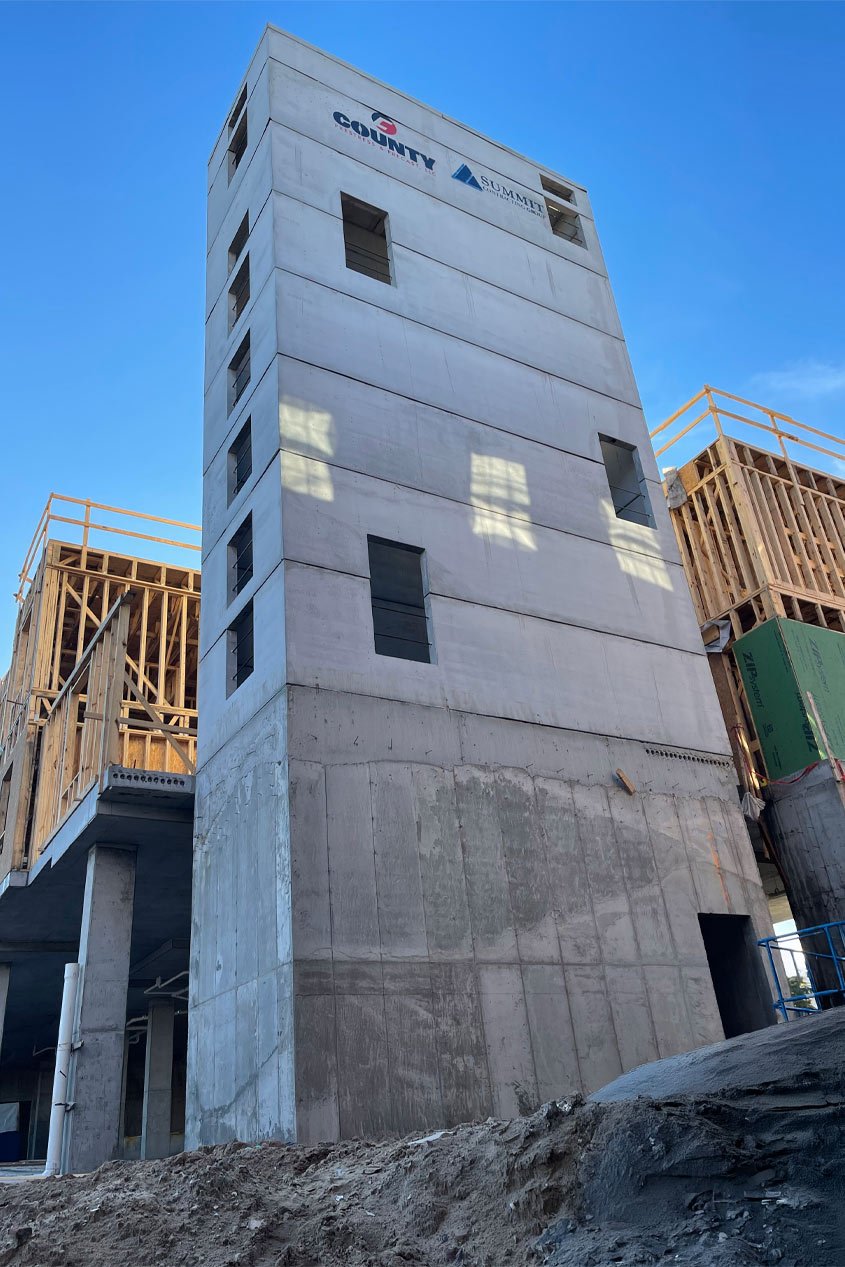 A four-story precast concrete stair shaft rises up from a concrete foundation, while the rest of the building is under construction around it.