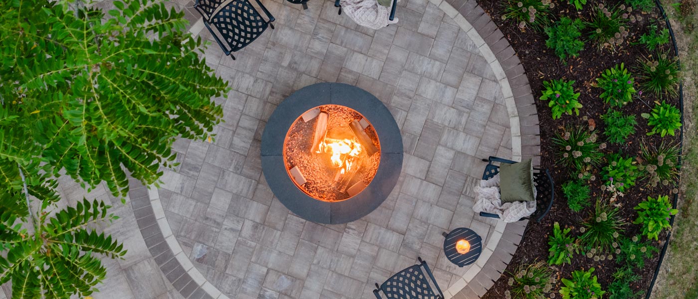 Overhead view of a circular paver patio surrounding a circular fire pit with a lit fire.