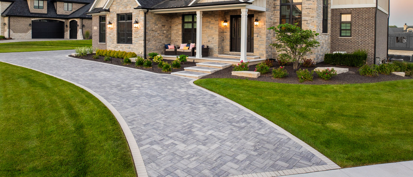 Gently curving gray paver driveway with contrasting border connecting to a low paver staircase in front of a large and elegant home.