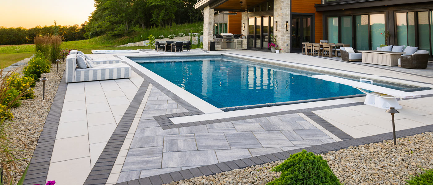 A swimming pool in front of a modern home with a concrete paver pool deck featuring a dramatic diagonal stripe made from three different paver colors